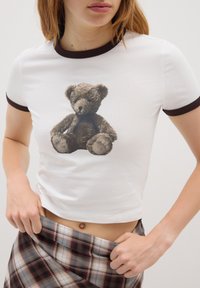 Camisa blanca de manga corta con ribete marrón que presenta un gráfico de un oso de peluche marrón sentado. El material parece suave y tiene un estiramiento moderado.