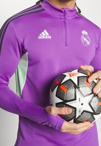 Lila träningsöverdel med långa ärmar och hög krage, med svarta och gröna detaljer. Håller en svartvit mönstrad fotboll med orange detaljer.