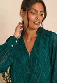 Blusa teal con scollo a V, chiusura con zip e motivo a onde testurizzato. Presenta maniche trasparenti, dettagli con bottoni e cuciture intricate.