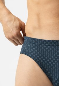 Hand die de zijkant van een donkerblauw gemusterd zwembriem aanpast op de heup en het onderste torso van een persoon tegen een witte achtergrond.