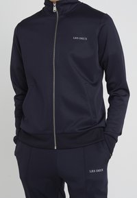 Muž v námořnickém modrém zipovém saka a odpovídajících kalhotách s logem "LES DEUX" na hrudi a stehně, s rukama v kapsách.