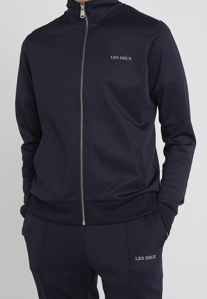 Muž v námořnickém modrém zipovém saka a odpovídajících kalhotách s logem "LES DEUX" na hrudi a stehně, s rukama v kapsách.