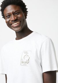 Weißes T-Shirt aus weichem Stoff, mit einem kleinen bestickten Design mit "Fishing Therapy" und einem Fisch. Kurze Ärmel und runder Kragen.