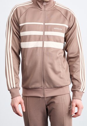 Chándal deportivo marrón con chaqueta de cremallera que presenta franjas crema y logo. Tejido suave con un diseño ajustado y puños acanalados.