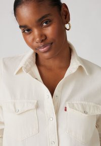 Camisa de mezclilla color crema con cierres a presión y dos bolsillos en el pecho. Presenta un cuello y una pequeña etiqueta roja de Levi's en el bolsillo izquierdo.