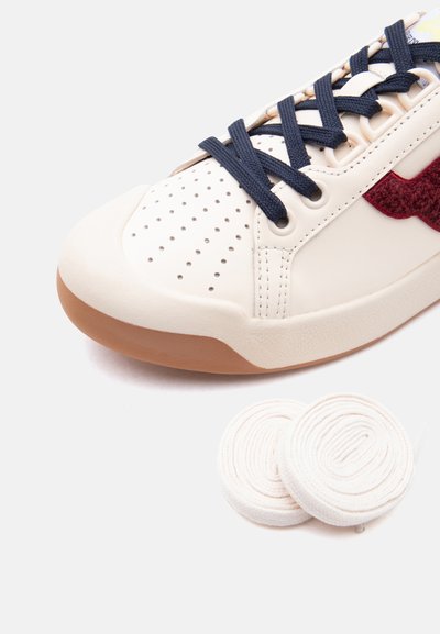 Baskets blanches avec lacets noirs, zone des orteils perforée, détail latéral texturé rouge, et deux lacets blancs roulés à côté sur un fond blanc.