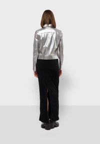 Veste métallique argentée au design ajusté, vue de dos, associée à une longue jupe noire avec des fentes sur les côtés et des bottes noires épaisses.