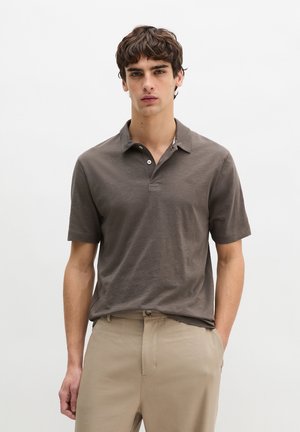 Jonge man met kort bruin haar, gekleed in een bruin poloshirt met korte mouwen, ingestopt in beige broek, staand tegen een effen lichte achtergrond.