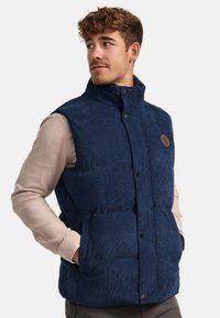 Marineblaues Steppwesten mit hohem Kragen, vorderen Druckknöpfen und seitlichen Taschen. Hergestellt aus strukturierter Stoff mit einem Logo-Patch auf der Brust.