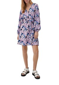 Robe florale violette avec un décolleté en V, de longues manches bouffantes et une taille ceinturée. Portée avec des sandales blanches à boucles et semelles noires.