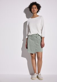 Helle grüne Jeansrock mit Wickeldesign und Knopfdetails, kombiniert mit einem lockeren weißen Langarmoberteil und hellen Sneakers.