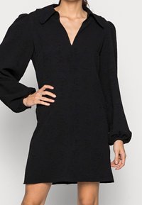 Kvinna som bär en strukturerad svart klänning med en djup V-ringning och långa puffärm, handen vilande på höften, stående mot en enfärgad bakgrund.