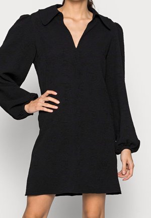 Donna che indossa un vestito nero texture con un profondo colletto a V e maniche lunghe gonfie, con la mano appoggiata sui fianchi, in piedi contro uno sfondo semplice.