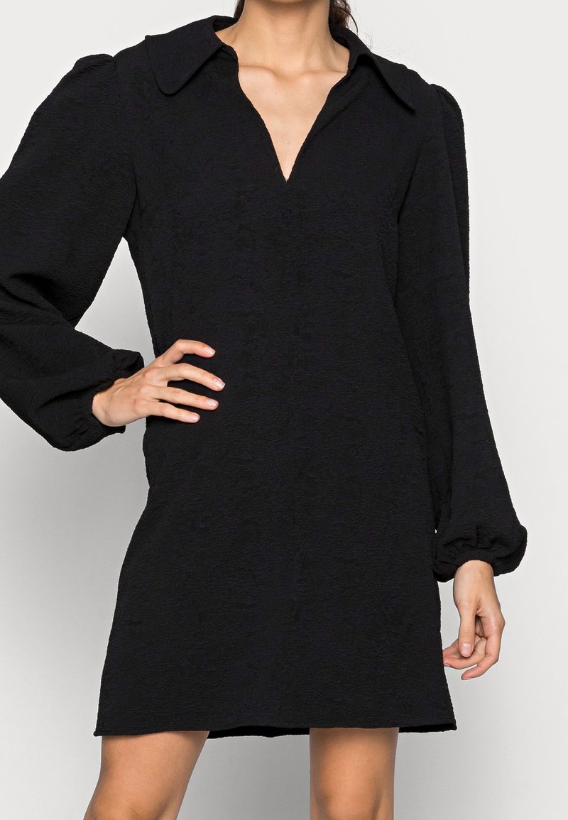 Kvinna som bär en strukturerad svart klänning med en djup V-ringning och långa puffärm, handen vilande på höften, stående mot en enfärgad bakgrund.