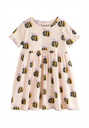 Kind trägt cremefarbenes Kleid mit schwarzen und gelben Bienenmuster, steht vor einer hellen strukturierten Wand, schaut nach links, lockiges Haar sichtbar.