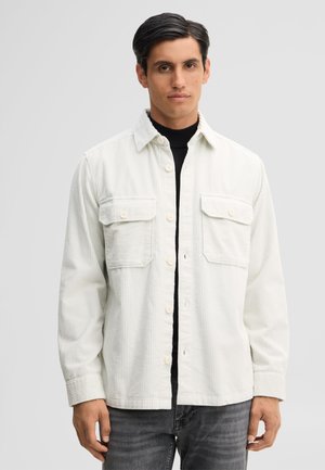 Mann mit dunklem Haar trägt ein weißes Cordhemd über einem schwarzen Rollkragen und ausgewaschene schwarze Jeans vor einem einfarbigen grauen Hintergrund.