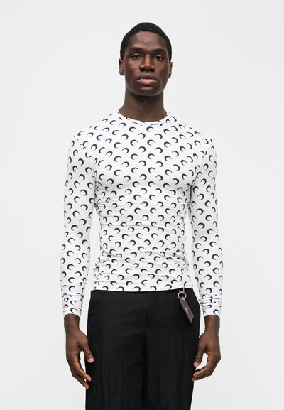 Man wearing tight white long-sleeve shirt with black crescent moon pattern and loose black pants, standing against plain light background.