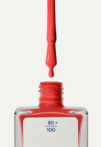 Lo smalto per unghie rosso cola da un pennello sopra una bottiglia di vetro trasparente con un'etichetta bianca che mostra "80 + 100" in testo blu.