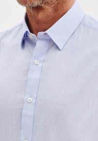 Jasnoniebieska koszula z guzikami, wykonana z teksturowanej tkaniny, z kołnierzykiem w szpic i białymi guzikami wzdłuż centralnego zapięcia.