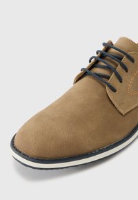 Brune snørebåndssko med en teksturert overflate, beige overdel, mørkeblå snøring, og en hvit såle. Merker seg sømdetaljer nær snøringer.