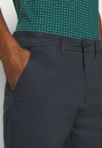 Marine chinos lavet af glat stof har en frontlomme og bæltestropper, parret med en grøn mønstret skjorte, der viser små firkantede designs.