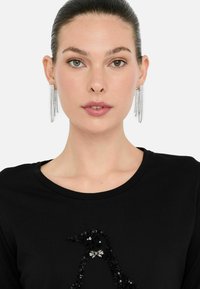T-shirt nera con un design di pinguino lucido realizzato in paillettes. Il modello indossa orecchini d'argento pendenti con più fili.