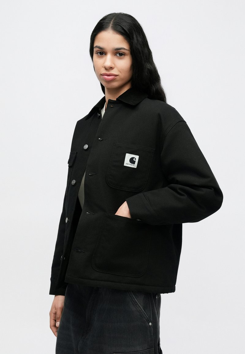 Joven con cabello largo y negro, vestido con una chaqueta negra Carhartt y la mano en el bolsillo, de pie frente a un fondo claro y liso.