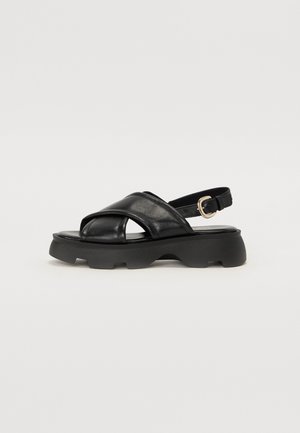 Black leather sandals with cross-strap design, adjustable ankle buckle, and chunky rubber sole. Smooth texture with a glossy finish.