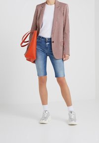 Blazer a cuadros rojos y blancos sobre una camiseta blanca, combinado con unos pantalones cortos de mezclilla azules y zapatillas plateadas. Se sostiene un bolso tote naranja brillante.