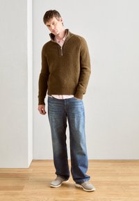 Brown knitted pullover with a zip collar, layered over a light pink shirt, paired with blue jeans and grey shoes, standing on wooden floor.
