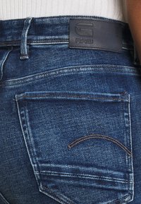 Jeans de mezclilla azul oscuro con una textura suave, diseño clásico de cinco bolsillos y una etiqueta de cuero visible con la inscripción "RAW" en la cinturilla.