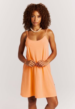 Vrouw met krullend haar, gekleed in een mouwloze oranje jurk en een witte kralenketting, staand met handen gevouwen voor zich.