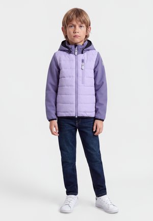 Kind met lichtbruin haar dat een lavendelkleurige gewatteerde jas met capuchon, donkere spijkerbroek en witte sneakers draagt, staand tegen een effen achtergrond.