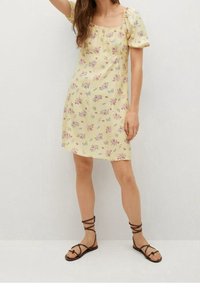 Vestido floral amarillo con mangas abullonadas y escote cuadrado, adornado con pequeñas flores rosas y azules. Combinado con sandalias marrones de tiras.