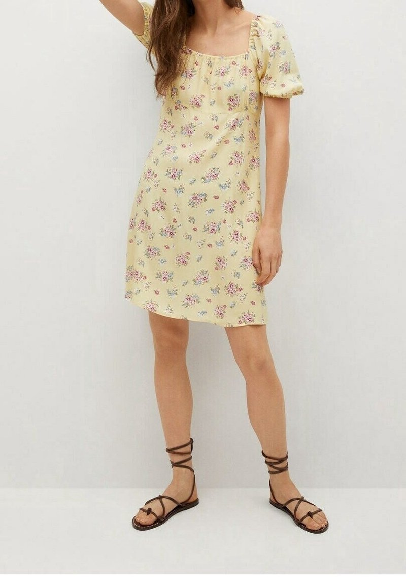 Vestido floral amarillo con mangas abullonadas y escote cuadrado, adornado con pequeñas flores rosas y azules. Combinado con sandalias marrones de tiras.