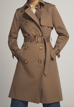 Woman wearing a beige double-breasted trench coat with belt and buckled straps on sleeves, hands in pockets, paired with blue jeans.
