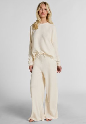 Vrouw staat blootsvoets, draagt een crèmekleurige gebreide top met lange mouwen en bijpassende wijduitlopende broek met trekkoord in de taille.