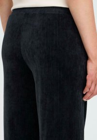 Donkere ribfluwelen broek met een hoge tailleband, voorzien van verticale lijnen en een gladde textuur. Zijzakken zijn niet zichtbaar in de afbeelding.