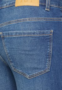 Jeans de denim azul com um patch de couro castanho, bolso traseiro proeminente com costura em contraste e um design justo que expõe a textura do denim.