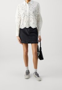 Blusa bianca in pizzo a occhio con orlo scallop, abbinata a una gonna di jeans nera e sneakers grigie. Una borsa nera aggiunge contrasto.