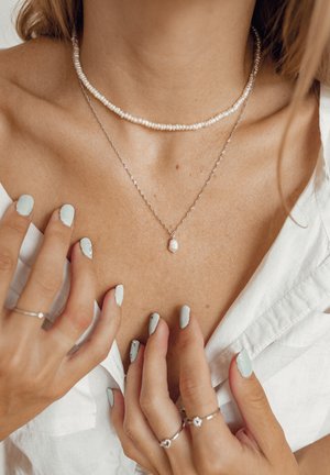 Layered pearl and silver necklaces on a bare neck, featuring a single pearl pendant. Hands with mint green nails touch the neckline.