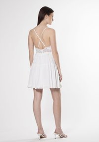 Vestido blanco acampanado con detalles de encaje, tirantes ajustables y dobladillo con volantes. Llevado con sandalias de tacón blancas.