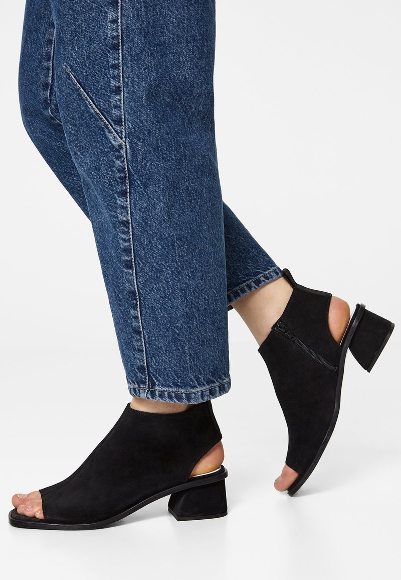 Chaussures à talons ouverts en daim noir avec un petit talon épais, fermeture éclair latérale et design découpé, associées à un jean en denim bleu.