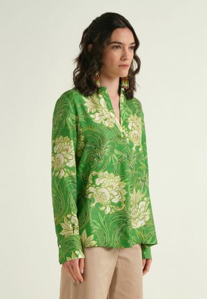 Femme portant un chemisier vert à motif floral avec des manches longues et un pantalon beige, légèrement tournée vers la droite sur un fond uni.