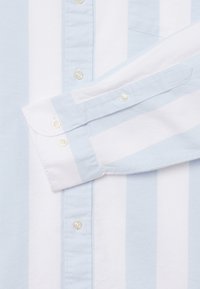 Poignet de chemise à manches longues à rayures verticales bleu clair et blanc, avec des boutons nacrés et un détail de patte de boutonnage boutonnée.