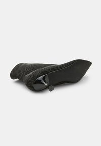 Zapato de tacón alto negro con un upper de malla texturizada, adornos sutiles y un tacón de charol brillante. Diseño de punta afilada.