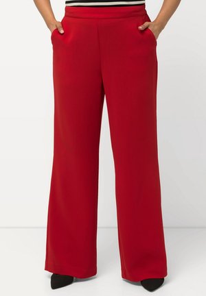 Personne portant un pantalon rouge taille haute à jambes larges, les mains dans les poches et des chaussures noires pointues visibles sous le pantalon.
