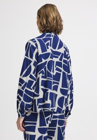 Blusa a maniche lunghe con motivo blu e beige, vestibilità morbida e polsini con bottone. Un design astratto copre il tessuto in varie forme.