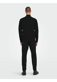 Suéter negro de punto con un cuello elevado, combinado con pantalones ajustados negros y zapatos negros, visto desde la espalda sobre un fondo claro.