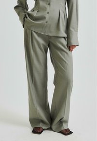 Pantaloni tailored de culoare gri deschis, confecționați dintr-un material neted, cu design evazat, având plissuri frontale și talie înaltă, asortați cu pantofi cu vârf deschis.